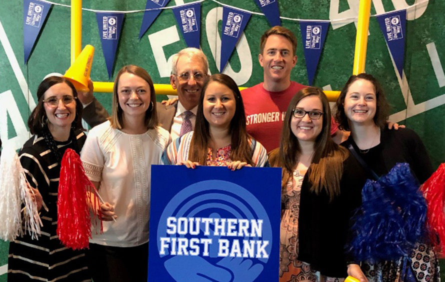 Group photo of 8 people in front of a football field backdrop and goal post, with one person in front holding up a Southern First Bank sign.