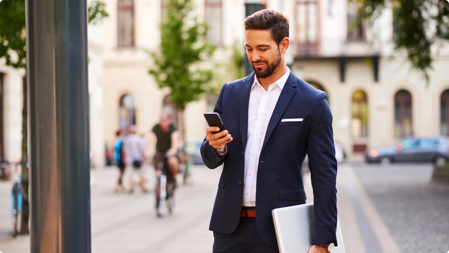 Business man with a laptop stopped on a sidewalk to check his phone, smiling, with a busy city scene in the background.