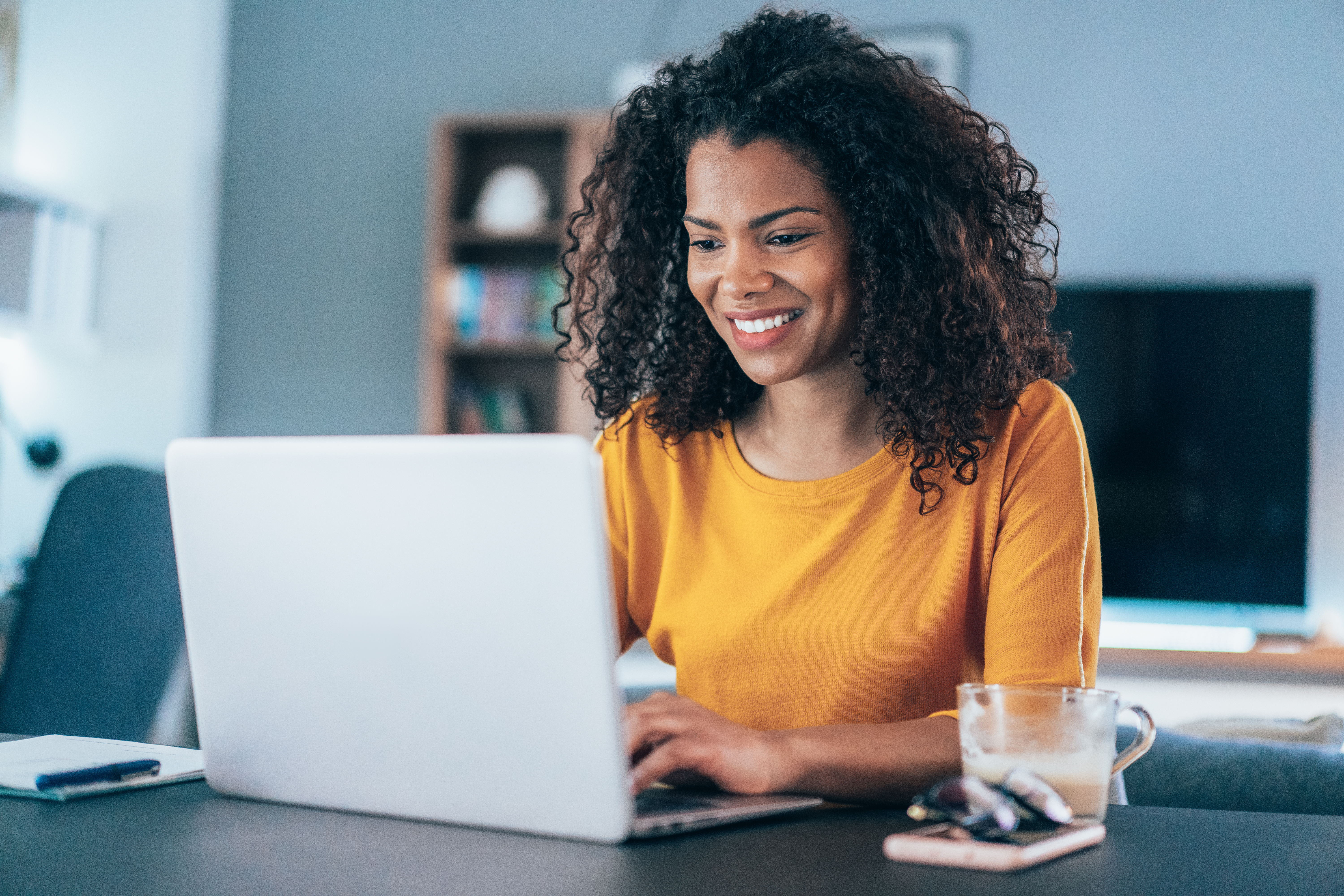 Woman with curly hair in gold shirt, smiling and looking at her laptop in front of her.
