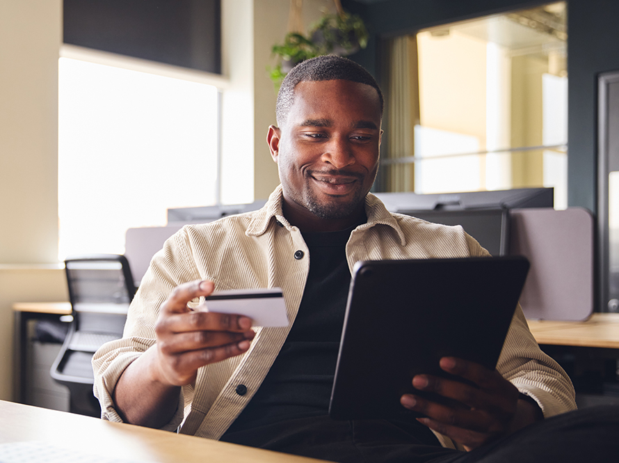 Man sitting at desk looking at credit card and tablet.
