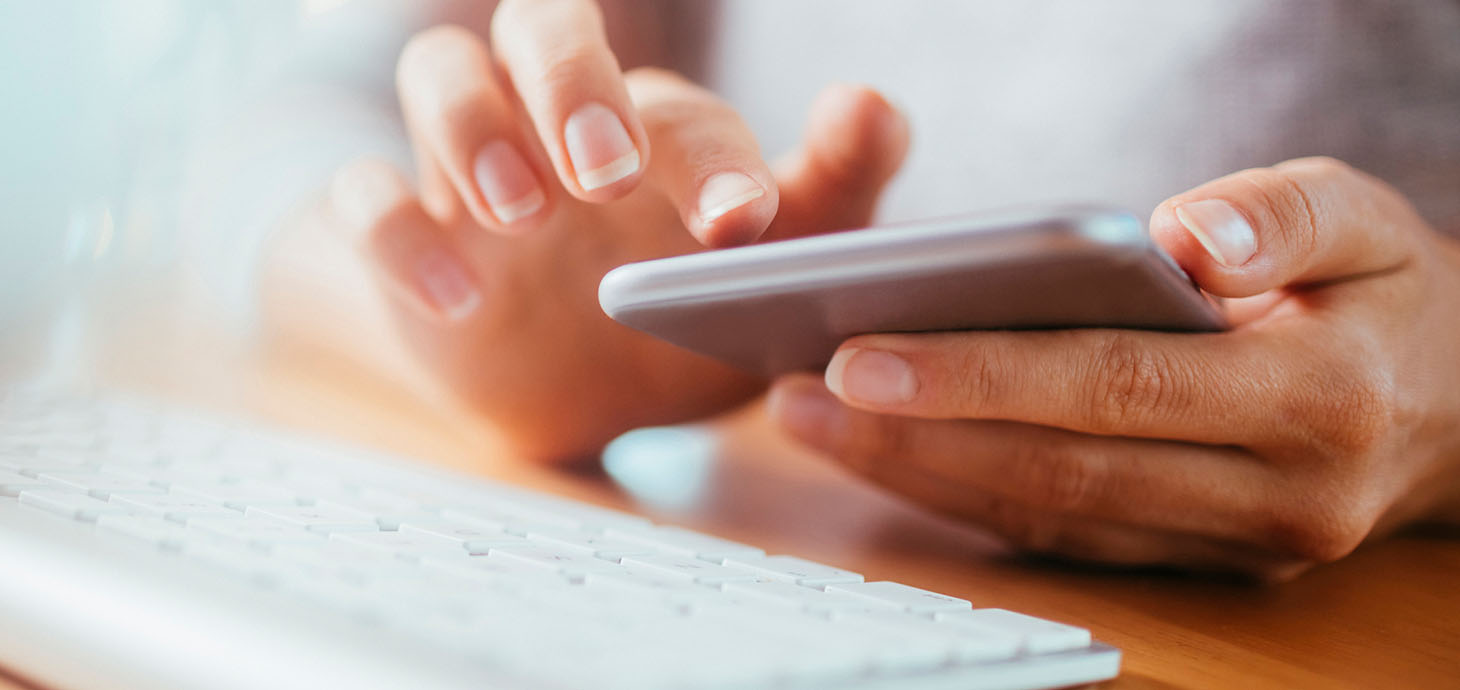 Hands tapping a smartphone being held over a keyboard.